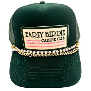May include: Dark green trucker hat with a mesh back and a curved brim. The front features a rectangular patch with the text "EARLY BIRDIE CADDIE CLUB" above pink stripes and small rhinestones. A gold chain with pearls accents the brim.