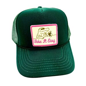 Pode incluir: Bon&eacute; de caminhoneiro verde escuro com parte traseira em malha e aba curva. A frente apresenta um patch retangular com um jacar&eacute; usando &oacute;culos de sol e segurando uma bebida, com o texto "Take It Easy" em rosa.