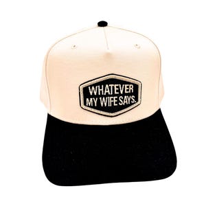 May include: A baseball cap with a cream-colored crown and a black brim. The cap features a black patch with the text "WHATEVER MY WIFE SAYS" in white lettering. The cap has a black button on top.