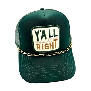 May include: A dark green trucker hat with a mesh back and a curved brim. The front patch reads "Y'ALL AIN'T RIGHT" in white, tan, and pink embroidery. A gold-colored chain is attached to the brim.