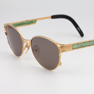 Op de afbeelding: Gouden Cartier zonnebril met groene accenten en bruine glazen. De zonnebril heeft een dun metalen frame en zwarte kunststof pootjes.