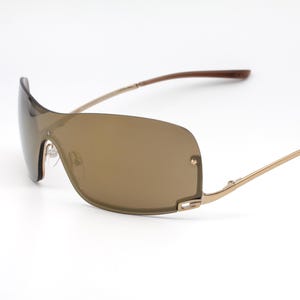 May include: Rimless sunglasses featuring a single continuous brown tinted lens that wraps around the front. The frame consists of thin gold-toned metal temples and a bridge, with dark brown plastic tips at the ends of the arms for a secure fit.