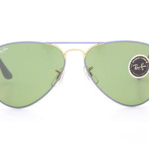 Op de afbeelding: Paarse en gouden aviator zonnebril met groene glazen. De zonnebril heeft een Ray-Ban logo op de linkerlens en een Ray-Ban sticker op de rechterlens.