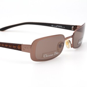 May include: Brown rectangular sunglasses with a light brown tint. The frames are a metallic brown color, with a textured pattern on the temples. The brand name "Christian Dior" is visible on the lens.