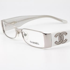 Può includere: Occhiali Chanel con montatura argentata e aste bianche. Le lenti sono rettangolari e le aste presentano il logo Chanel. La parola "CHANEL" è stampata sulla lente.
