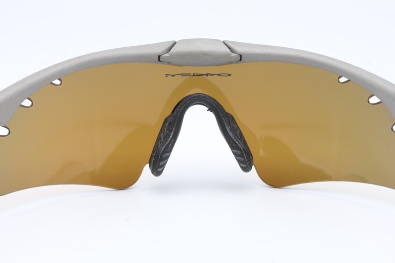 May include: Gray wraparound sunglasses with gold mirrored lenses. The sunglasses have a black nose bridge and black temple tips.
