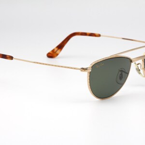 May include: Gold-framed sunglasses with dark green lenses. The sunglasses have a double bridge and tortoise-shell patterned temples. A small sticker with a logo is on the right lens. The glasses are set against a white background.
