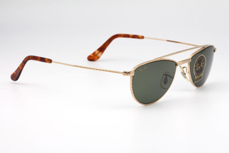 May include: Gold-framed sunglasses with dark green lenses. The sunglasses have a double bridge and tortoise-shell patterned temples. A small sticker with a logo is on the right lens. The glasses are set against a white background.