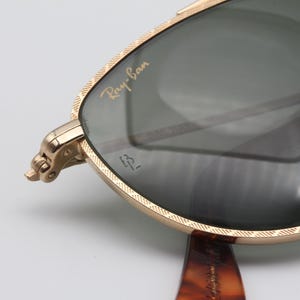 May include: Close-up of Ray-Ban sunglasses with a gold-colored frame and dark lenses. The Ray-Ban logo is visible on the lens. The frame has a textured design. The temples are a tortoiseshell pattern.