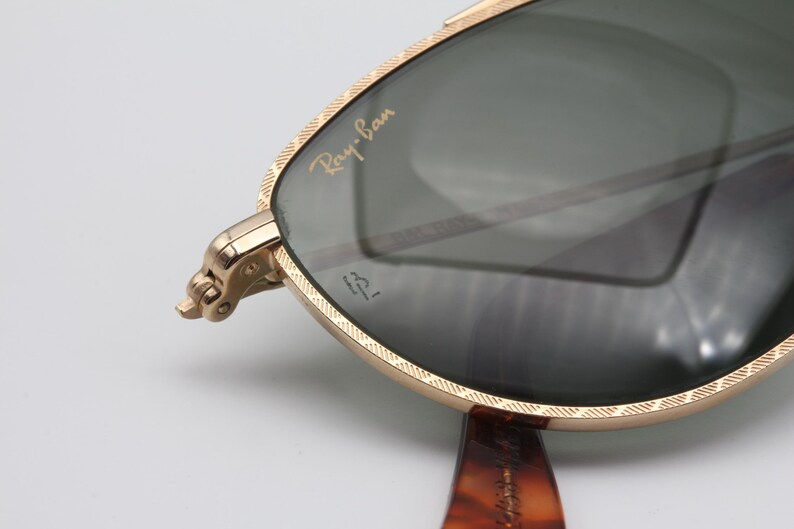 May include: Close-up of Ray-Ban sunglasses with a gold-colored frame and dark lenses. The Ray-Ban logo is visible on the lens. The frame has a textured design. The temples are a tortoiseshell pattern.