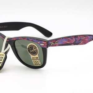 May include: Black Ray-Ban Wayfarer sunglasses with a purple and pink camouflage pattern on the arms. The lenses are green and the sunglasses have a gold Ray-Ban logo on the right lens.