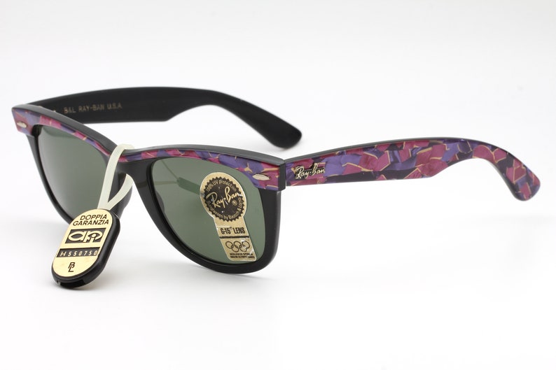 May include: Black Ray-Ban Wayfarer sunglasses with a purple and pink camouflage pattern on the arms. The lenses are green and the sunglasses have a gold Ray-Ban logo on the right lens.