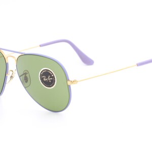 Op de afbeelding: Paarse en gouden aviator zonnebril met groene glazen. De zonnebril heeft een Ray-Ban logo op de rechter lens.