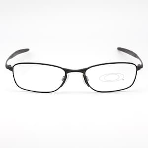 Op de afbeelding: Zwarte rechthoekige bril met heldere glazen. De bril heeft een dun, donker montuur en een subtiele neusbrug. Het Oakley-logo is zichtbaar op de rechterlens. De pootjes buigen lichtjes.