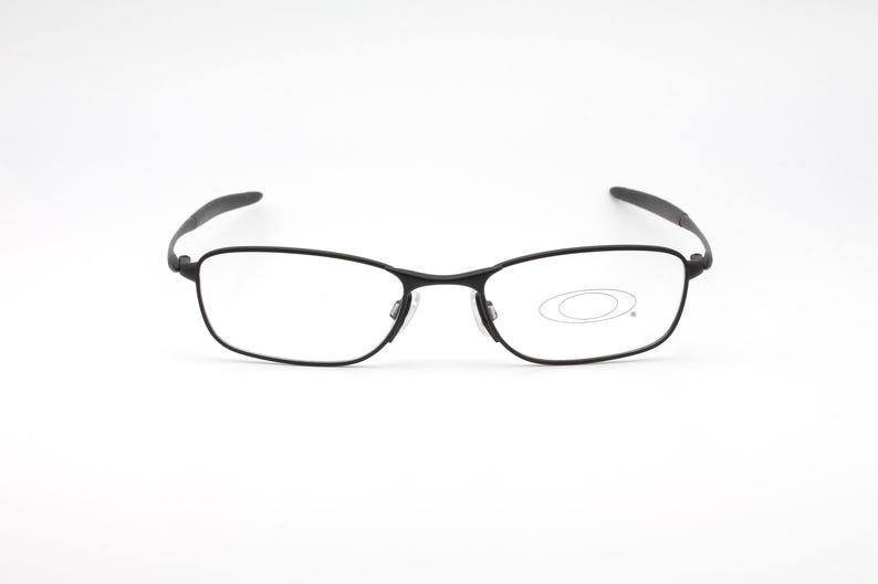 Op de afbeelding: Zwarte rechthoekige bril met heldere glazen. De bril heeft een dun, donker montuur en een subtiele neusbrug. Het Oakley-logo is zichtbaar op de rechterlens. De pootjes buigen lichtjes.