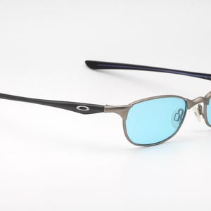 May include: A pair of Oakley eyeglasses with a gunmetal frame and blue lenses. The temples are black with the Oakley logo. The lenses are a bright blue color.