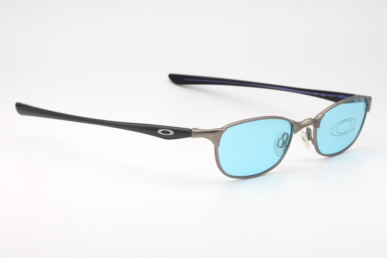 May include: A pair of Oakley eyeglasses with a gunmetal frame and blue lenses. The temples are black with the Oakley logo. The lenses are a bright blue color.