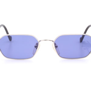 Può includere: Occhiali da sole argentati con montatura rettangolare e lenti blu. Gli occhiali hanno un dettaglio decorativo sulla cerniera.