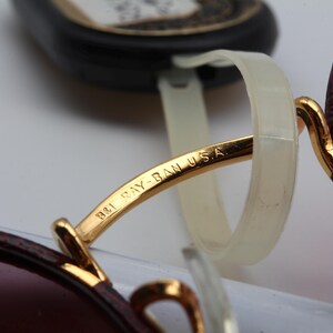 May include: Close-up of a pair of gold-rimmed sunglasses with a brown leather frame. The sunglasses have a pink lens and the words "B&L Ray-Ban USA" are engraved on the frame.