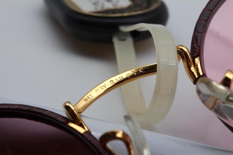 May include: Close-up of a pair of gold-rimmed sunglasses with a brown leather frame. The sunglasses have a pink lens and the words "B&L Ray-Ban USA" are engraved on the frame.