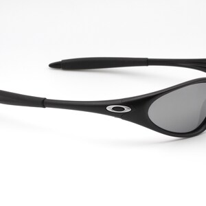 May include: Black sunglasses with gray lenses. The sunglasses have a curved frame and arms, with a silver logo on the side. The lenses are a dark gray color, and the frame is a matte black.