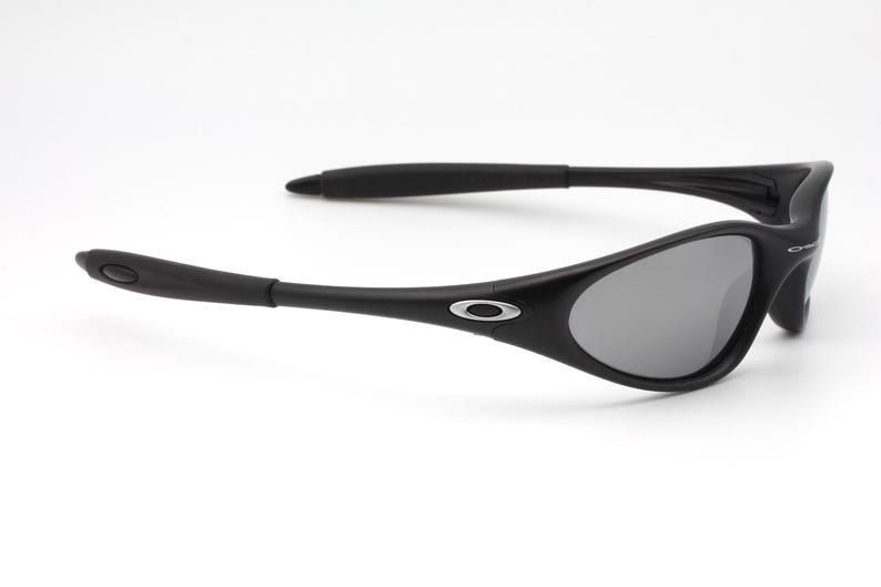 May include: Black sunglasses with gray lenses. The sunglasses have a curved frame and arms, with a silver logo on the side. The lenses are a dark gray color, and the frame is a matte black.