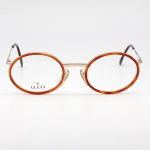May include: Oval-shaped eyeglasses featuring a tortoiseshell-patterned frame with a gold-tone metal bridge and thin temples. The left lens displays the Gucci brand name and logo in black text. These glasses have clear lenses and adjustable nose pads.