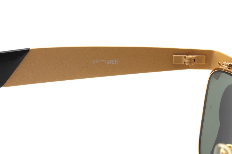 Puede incluir: Gafas de sol Wayfarer de color dorado con montura negra y lentes verdes. Las gafas est&aacute;n dobladas por la mitad y el logotipo de Wayfarer es visible en la patilla.