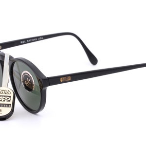 May include: Black round Ray-Ban sunglasses with green lenses. The sunglasses have a gold Ray-Ban logo on the side. The sunglasses are accompanied by a black tag with the text "Doppia Garanzia" and a serial number.