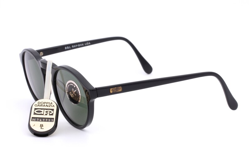 May include: Black round Ray-Ban sunglasses with green lenses. The sunglasses have a gold Ray-Ban logo on the side. The sunglasses are accompanied by a black tag with the text "Doppia Garanzia" and a serial number.