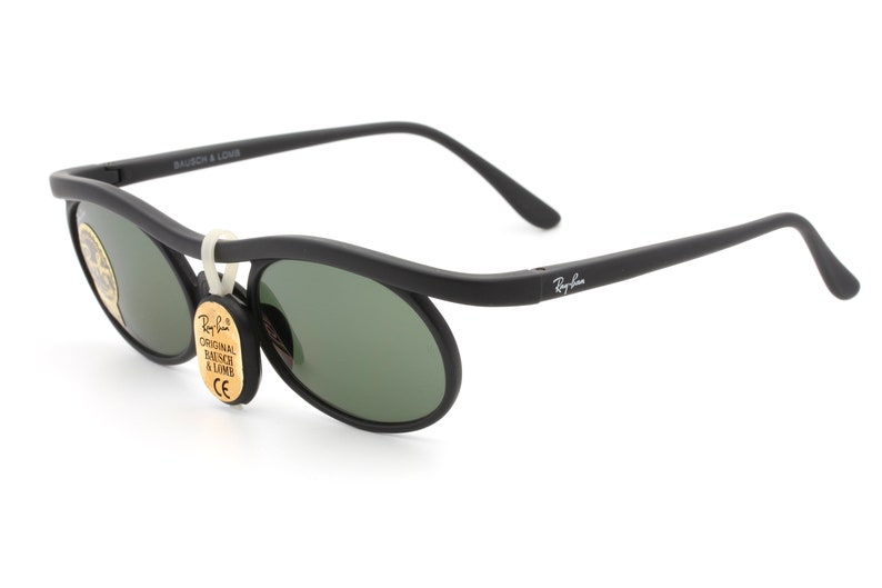 May include: Black Ray-Ban sunglasses with green lenses and a curved frame. The sunglasses have a tag that reads "Original Bausch & Lomb CE".