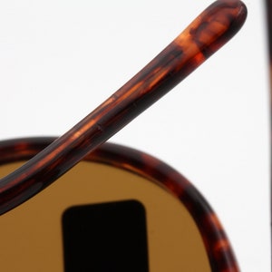 May include: Close-up of sunglasses with a tortoiseshell pattern on the frames. The lenses are a light brown color. The sunglasses are set against a white background.