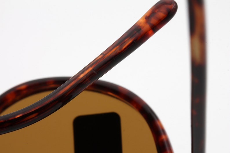 May include: Close-up of sunglasses with a tortoiseshell pattern on the frames. The lenses are a light brown color. The sunglasses are set against a white background.