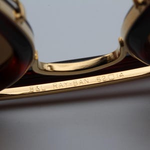 May include: Close-up of Ray-Ban sunglasses with gold frames and brown lenses. The text "B&L RAY-BAN 62 14" is engraved on the gold frame. The sunglasses have a classic design.