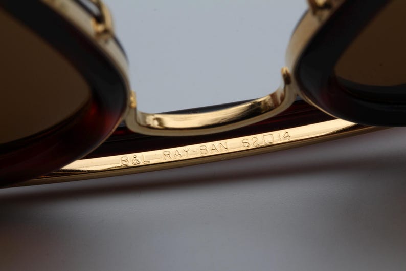 May include: Close-up of Ray-Ban sunglasses with gold frames and brown lenses. The text "B&L RAY-BAN 62 14" is engraved on the gold frame. The sunglasses have a classic design.