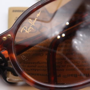 May include: Close-up of a pair of brown tortoise shell Ray-Ban sunglasses with gold lettering on the frame. The sunglasses are reflecting light and a blurry image of text.