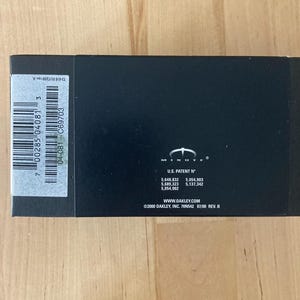 May include: A black rectangular box with a white barcode label and the text "7 00285 04081 3". The box also features the Oakley logo and patent numbers. The text "WWW.OAKLEY.COM" is visible.