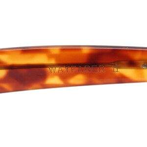 May include: Close-up of the arm of Wayfarer II sunglasses. The frame is a tortoiseshell pattern in shades of brown and amber. The text "WAYFARER II" is visible on the arm.