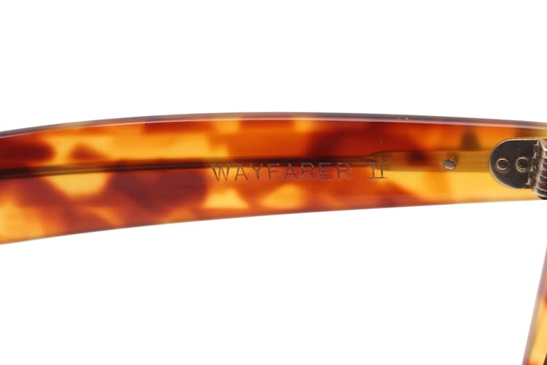 May include: Close-up of the arm of Wayfarer II sunglasses. The frame is a tortoiseshell pattern in shades of brown and amber. The text "WAYFARER II" is visible on the arm.