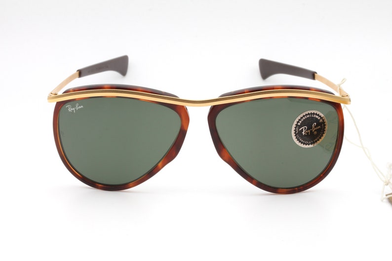 Pu&ograve; includere: Occhiali da sole con montatura tartaruga, dettagli dorati e lenti verdi. Gli occhiali hanno il logo Ray-Ban sulla lente sinistra e un adesivo Ray-Ban sulla lente destra.