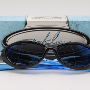 May include: Black sunglasses with blue lenses and a blue frame. The sunglasses are on a blue and white box with the word "Oakley" written on it.