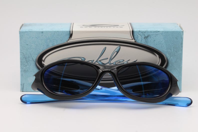 May include: Black sunglasses with blue lenses and a blue frame. The sunglasses are on a blue and white box with the word "Oakley" written on it.