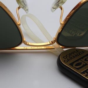 May include: Gold-tone aviator sunglasses with green lenses. The sunglasses have a Ray-Ban logo on the side and a black tag with the text "Doppia Garanzia" and a serial number.