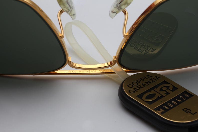 May include: Gold-tone aviator sunglasses with green lenses. The sunglasses have a Ray-Ban logo on the side and a black tag with the text "Doppia Garanzia" and a serial number.