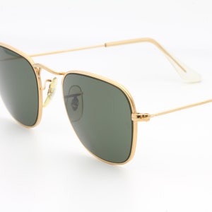May include: Gold-framed square sunglasses with green lenses. The sunglasses are on a white background.