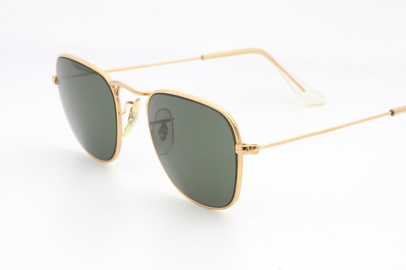 May include: Gold-framed square sunglasses with green lenses. The sunglasses are on a white background.