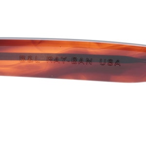 May include: Close-up of the brown tortoiseshell frame of a pair of Ray-Ban sunglasses. The text "B&L RAY-BAN USA" is engraved on the frame.