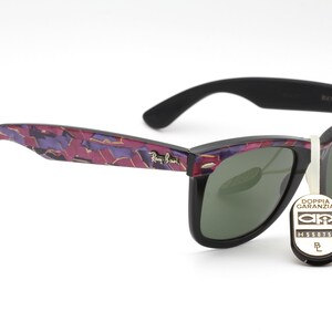 May include: Black Ray-Ban Wayfarer sunglasses with a purple and gold camouflage pattern on the arms. The lenses are green and the sunglasses have a tag that says "Doppia Garanzia" with a logo and the number "H558758".