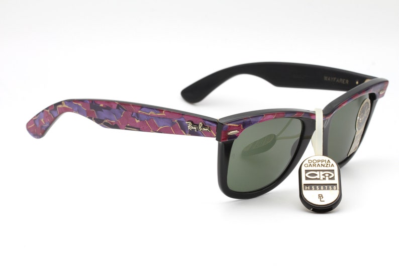 May include: Black Ray-Ban Wayfarer sunglasses with a purple and gold camouflage pattern on the arms. The lenses are green and the sunglasses have a tag that says "Doppia Garanzia" with a logo and the number "H558758".