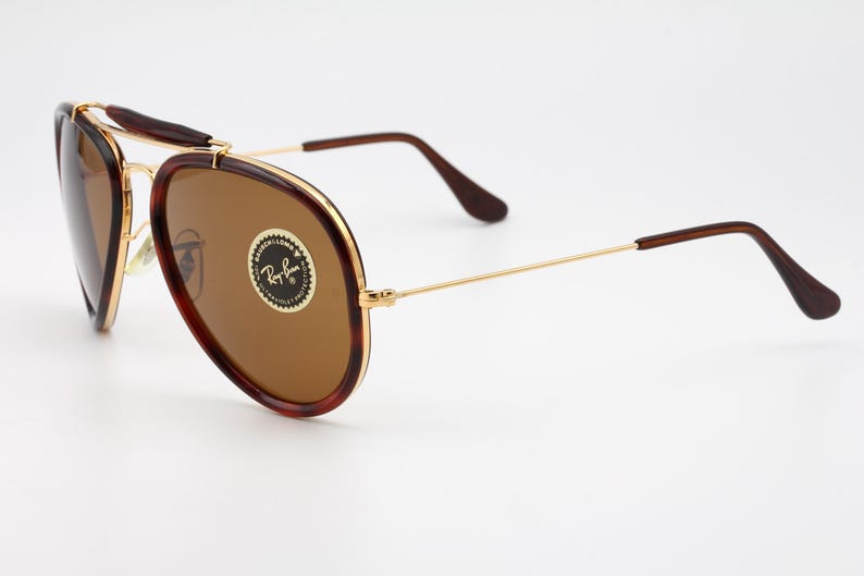 May include: Aviator-style sunglasses with brown lenses and a gold frame. The frames have a tortoiseshell pattern on the top and sides. The Ray-Ban logo is visible on the lens.